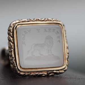 May include: A square, gold-toned ring with a clear, engraved stone featuring a lion and text. The ring has a decorative, textured border. The lion is standing on a small patch of ground.
