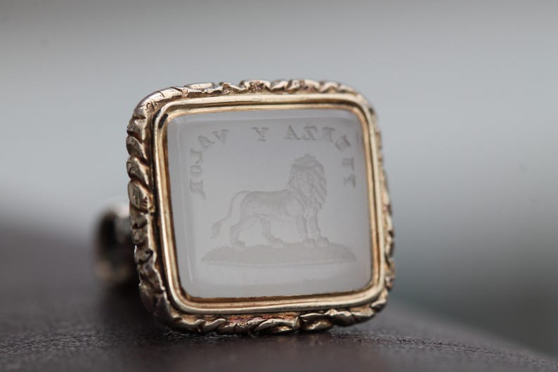 May include: A square, gold-toned ring with a clear, engraved stone featuring a lion and text. The ring has a decorative, textured border. The lion is standing on a small patch of ground.