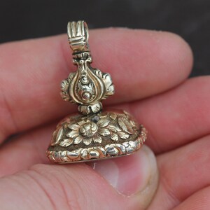 May include: A detailed, antique gold-colored seal with floral and ornate designs. The seal has a loop at the top and is held in a hand. The bottom of the seal features a floral pattern.