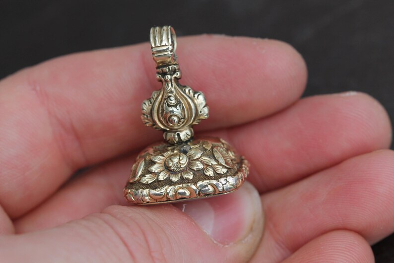 May include: A detailed, antique gold-colored seal with floral and ornate designs. The seal has a loop at the top and is held in a hand. The bottom of the seal features a floral pattern.