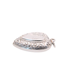 May include: A silver heart-shaped pendant with ornate scrollwork and a blank, rectangular space for engraving. The pendant has a small loop for attaching to a chain. The overall design is detailed and elegant.