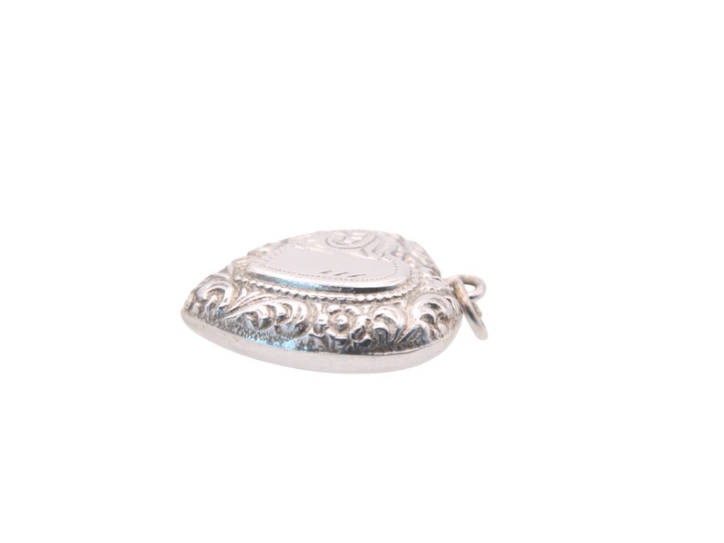 May include: A silver heart-shaped pendant with ornate scrollwork and a blank, rectangular space for engraving. The pendant has a small loop for attaching to a chain. The overall design is detailed and elegant.