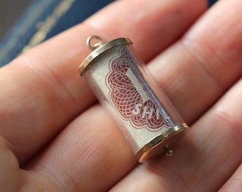 Può includere: Un piccolo ciondolo cilindrico con cappucci color oro e un corpo in vetro trasparente. All'interno, un foglio di carta arrotolato con un disegno rosso e bianco e il numero "1 SHI" è visibile. Il ciondolo è tenuto in una mano.