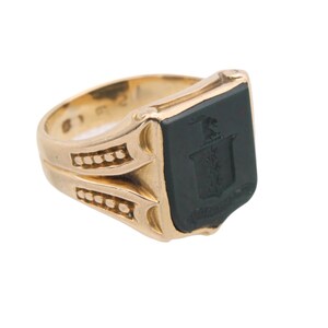May include: A gold signet ring featuring a flat, rectangular black onyx inlay. The centre of the black stone displays an engraved heraldic crest or coat of arms. The gold band has decorative side detailing with small, raised circular accents.