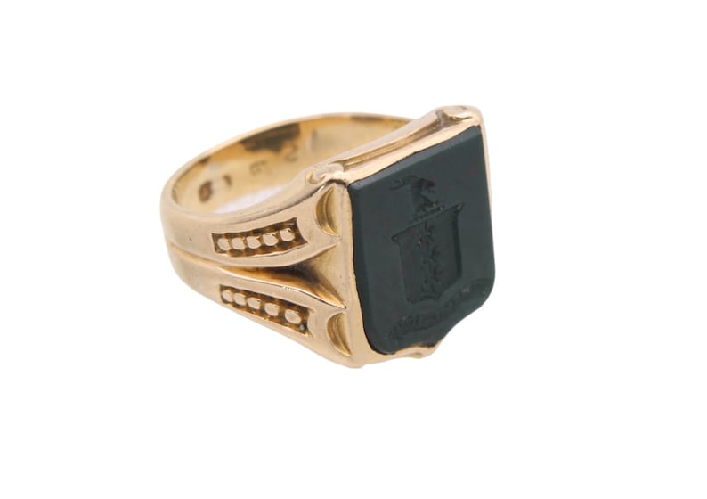 May include: A gold signet ring featuring a flat, rectangular black onyx inlay. The centre of the black stone displays an engraved heraldic crest or coat of arms. The gold band has decorative side detailing with small, raised circular accents.