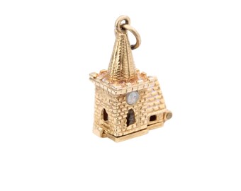 Puede incluir: Un charm dorado con forma de iglesia con campanario. El charm presenta un detallado patrón de ladrillos y pequeñas ventanas. Un pequeño lazo en la parte superior para sujetarlo a una pulsera o collar. El charm está sobre un fondo blanco.