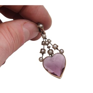 Puede incluir: Un colgante vintage con una piedra preciosa en forma de coraz&oacute;n, de color p&uacute;rpura claro. El colgante est&aacute; adornado con peque&ntilde;as perlas y un marco de metal dorado. El dise&ntilde;o incluye una serie de perlas suspendidas sobre el coraz&oacute;n, creando un aspecto delicado y elegante.