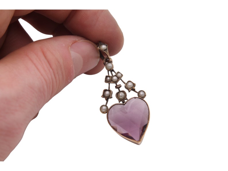 Puede incluir: Un colgante vintage con una piedra preciosa en forma de coraz&oacute;n, de color p&uacute;rpura claro. El colgante est&aacute; adornado con peque&ntilde;as perlas y un marco de metal dorado. El dise&ntilde;o incluye una serie de perlas suspendidas sobre el coraz&oacute;n, creando un aspecto delicado y elegante.