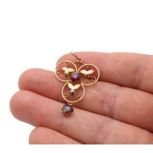 May include: A gold pendant with a floral design, featuring three red gemstones and a dangling single gemstone. The pendant is held in a hand against a white background. The pendant's design includes circular and leaf-like elements.