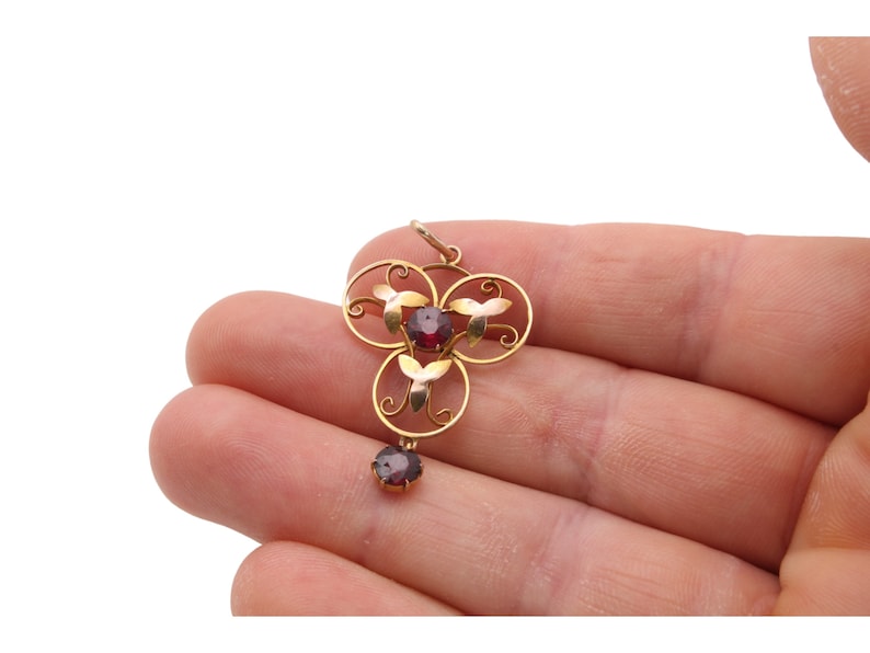 May include: A gold pendant with a floral design, featuring three red gemstones and a dangling single gemstone. The pendant is held in a hand against a white background. The pendant's design includes circular and leaf-like elements.