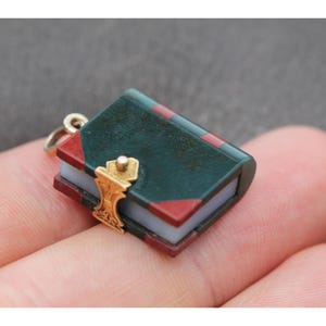 May include: Miniature book charm with a gold clasp. The book has a dark green cover with red accents and a gold-colored clasp. The pages are a light blue color. The charm is attached to a small gold-colored ring.