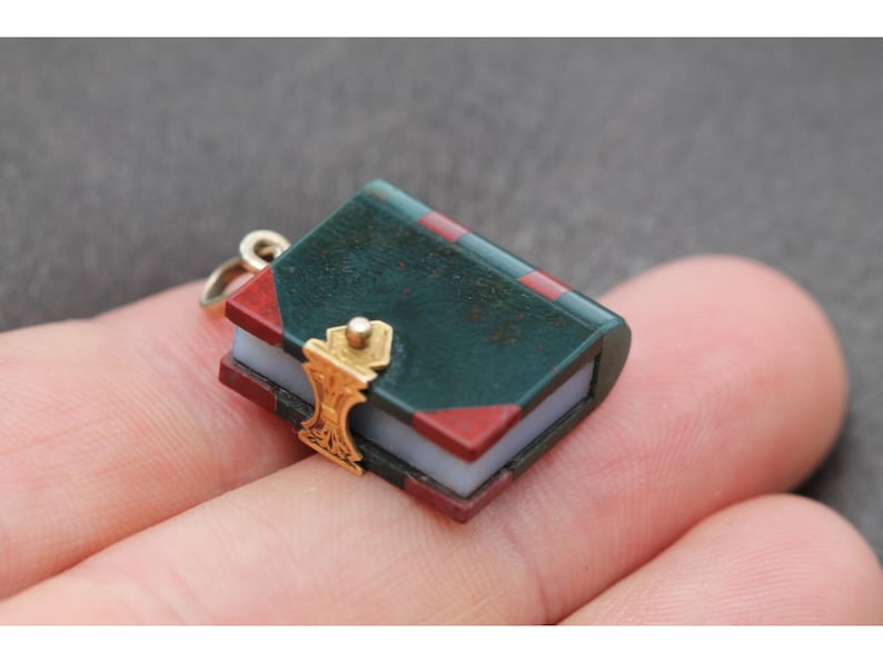 May include: Miniature book charm with a gold clasp. The book has a dark green cover with red accents and a gold-colored clasp. The pages are a light blue color. The charm is attached to a small gold-colored ring.