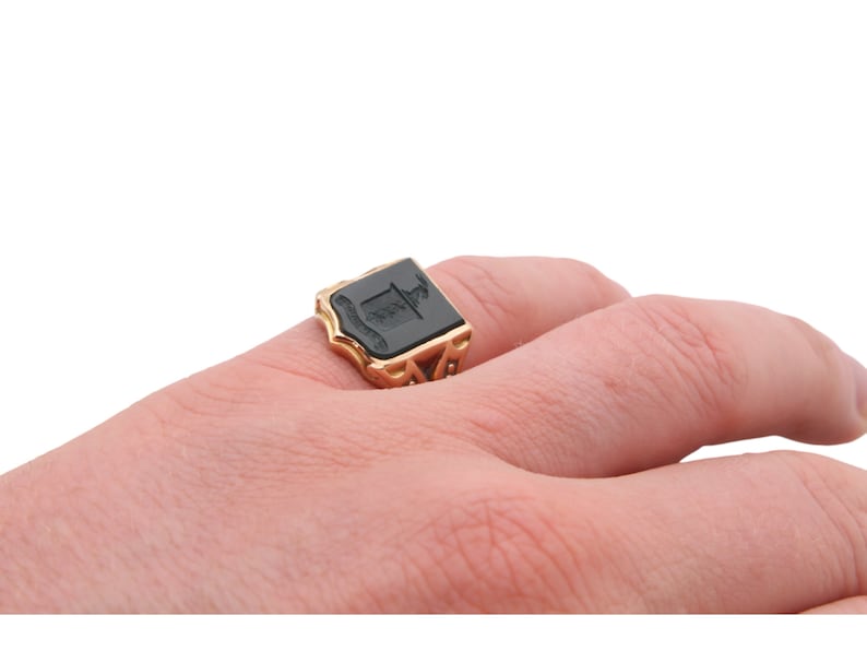 May include: A gold signet ring featuring a rectangular black onyx inlay with an engraved crest design. The metal band has an intricate openwork pattern along the sides, supporting the flat, dark stone face that sits prominently on the finger.