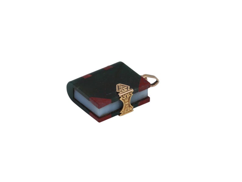 May include: A miniature book charm with a dark green cover and red corner accents. The book has a gold-colored clasp and a small loop for attaching to a chain or bracelet. The charm is set against a white background.