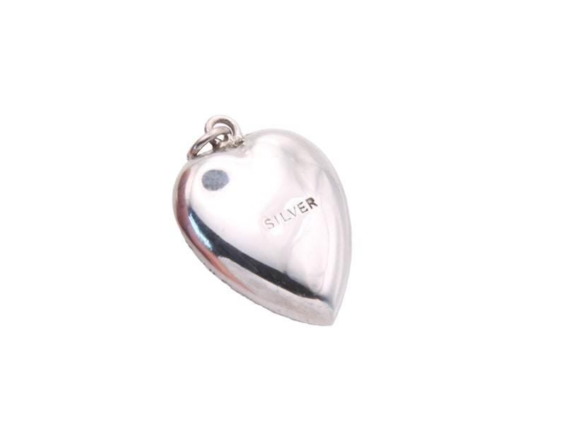 May include: A polished silver heart-shaped pendant with a small loop at the top for a chain. The word "SILVER" is stamped on the back of the pendant. The pendant is smooth and reflective.