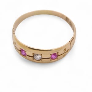 May include: A gold ring with three gemstones. The ring features a central clear gemstone flanked by two pink gemstones. The band has decorative etchings and is set against a white background. Jewelry.