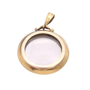 Op de afbeelding: Een goudkleurige, ronde medaillonhanger met een helder venster van glas of plastic. De hanger heeft een kleine lus aan de bovenkant voor een ketting en een decoratieve beugel. De medaillon is waarschijnlijk van metaal.
