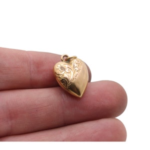 May include: A gold heart-shaped pendant with an ornate, engraved design on the front. The pendant has a small loop at the top for a chain. The heart is smooth and polished, reflecting light. The pendant is held in a person's hand.