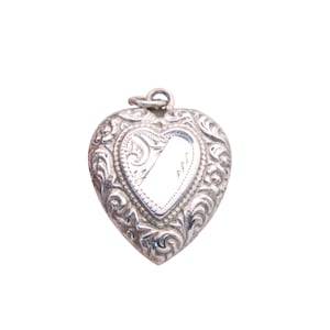 May include: A silver heart-shaped locket with ornate floral scrollwork. The locket has a small loop at the top for a chain and a blank, reflective center. The locket is a vintage style and is likely made of sterling silver.