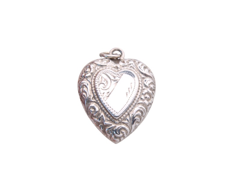 May include: A silver heart-shaped locket with ornate floral scrollwork. The locket has a small loop at the top for a chain and a blank, reflective center. The locket is a vintage style and is likely made of sterling silver.