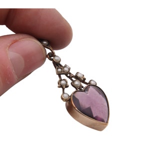 Puede incluir: Un colgante vintage con una piedra preciosa de amatista en forma de coraz&oacute;n. El colgante, con marco dorado, est&aacute; adornado con peque&ntilde;as perlas. El dise&ntilde;o incluye una parte superior decorativa con acentos de perlas adicionales, creando una joya elegante y atemporal.