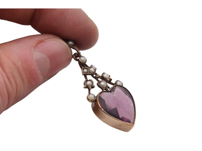 Puede incluir: Un colgante vintage con una piedra preciosa de amatista en forma de coraz&oacute;n. El colgante, con marco dorado, est&aacute; adornado con peque&ntilde;as perlas. El dise&ntilde;o incluye una parte superior decorativa con acentos de perlas adicionales, creando una joya elegante y atemporal.