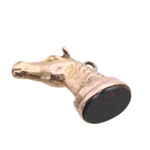 May include: A vintage gold-toned horse head seal with a dark red and black stone base. The horse head is detailed with a loop at the top. The seal is set against a white background.