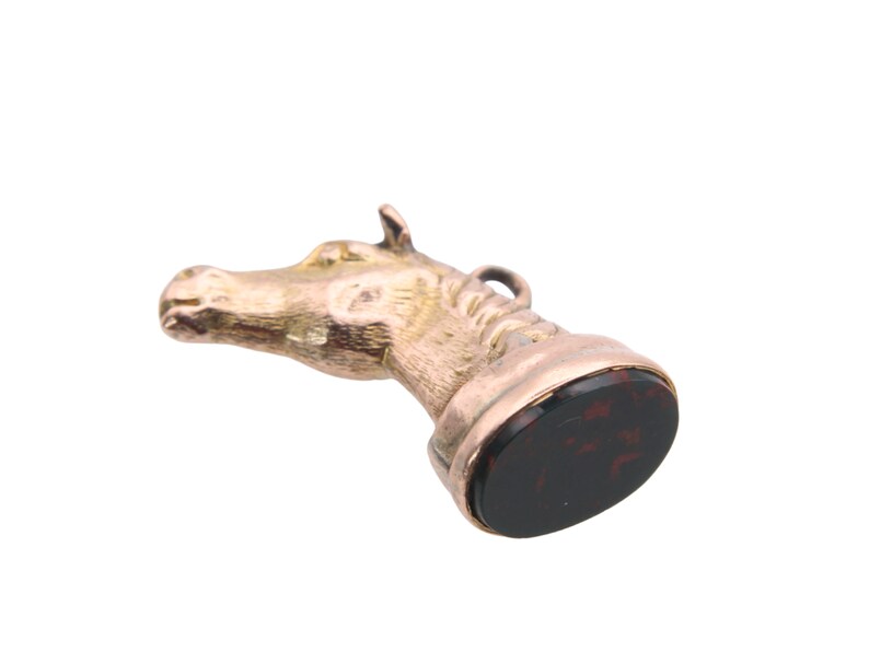 May include: A vintage gold-toned horse head seal with a dark red and black stone base. The horse head is detailed with a loop at the top. The seal is set against a white background.