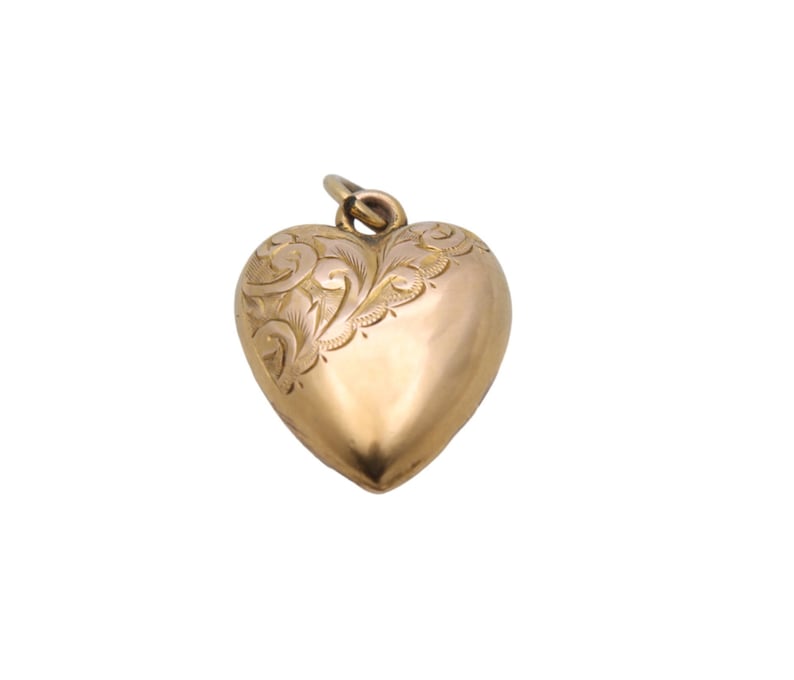 May include: A gold-colored heart-shaped pendant with an ornate, engraved floral design on one side. The pendant has a rounded, three-dimensional form and a small loop at the top for a chain. The overall aesthetic is vintage or antique.