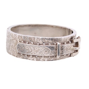 Pode incluir: Uma pulseira de bracelete articulada prateada com fecho de fivela. A pulseira apresenta um padrão floral gravado em sua superfície. A fivela e a placa retangular também são decoradas com intrincados desenhos.