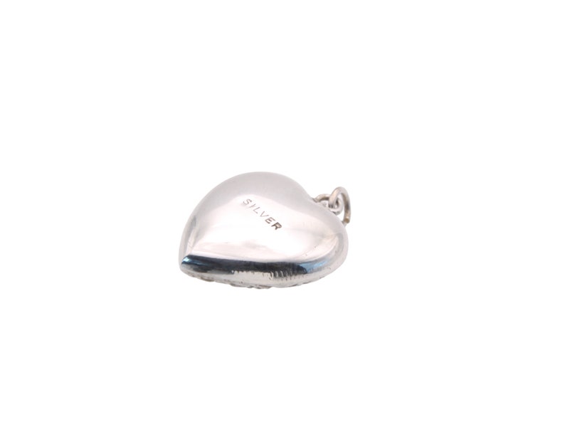 May include: A silver heart-shaped charm with a small loop at the top for attachment. The word "SILVER" is stamped on the surface of the charm. The charm is smooth and reflective, with a rounded shape.
