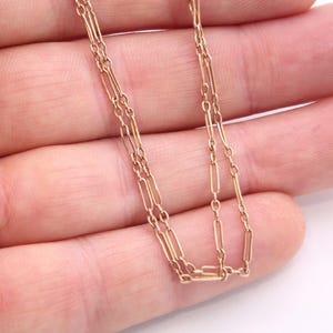 K&ouml;nnte beinhalten: Eine zarte Goldkette mit einem doppelreihigen Design. Die Halskette hat kleine, rechteckige Glieder, die zur Kette verbunden sind. Die warme Goldfarbe kontrastiert mit dem Hintergrund.
