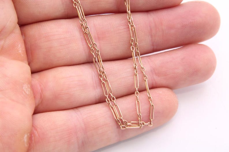K&ouml;nnte beinhalten: Eine zarte Goldkette mit einem doppelreihigen Design. Die Halskette hat kleine, rechteckige Glieder, die zur Kette verbunden sind. Die warme Goldfarbe kontrastiert mit dem Hintergrund.