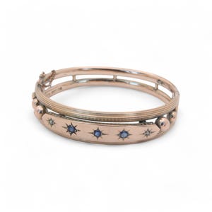 Op de afbeelding: Een roodgouden scharnierende armband. De armband heeft een reeks blauwe stervormige edelstenen en kleine diamantaccenten. De armband heeft een decoratief ontwerp met een sluiting.
