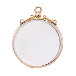 Op de afbeelding: Een goudkleurige, ronde hanger met een helder, cirkelvormig glazen midden. De hanger heeft een kleine lus aan de bovenkant om op te hangen, waarschijnlijk een medaillon of bedel. Het ontwerp is eenvoudig en elegant.