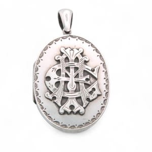 May include: Oval silver locket pendant with a decorative bail. The front features an ornate monogram design within a detailed border. The locket is a classic piece of jewelry, suitable for holding a photograph or small memento.