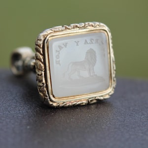 May include: A gold-toned signet ring with a square face. The face features a lion engraving and text. The ring has a twisted rope design around the edges. The background is blurred.