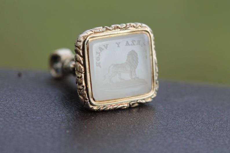 May include: A gold-toned signet ring with a square face. The face features a lion engraving and text. The ring has a twisted rope design around the edges. The background is blurred.