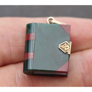 May include: Miniature book pendant with a gold clasp and bail. The book has a dark green cover with red accents and a gold clasp. The pendant is small and detailed, suitable for a necklace or charm bracelet.