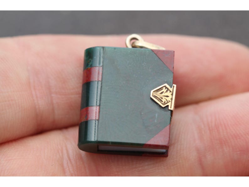 May include: Miniature book pendant with a gold clasp and bail. The book has a dark green cover with red accents and a gold clasp. The pendant is small and detailed, suitable for a necklace or charm bracelet.