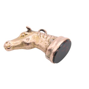 May include: A gold-colored horse head pendant with a dark red, circular stone base. The horse head is detailed with a textured mane and facial features. The pendant has a small loop at the top for hanging.