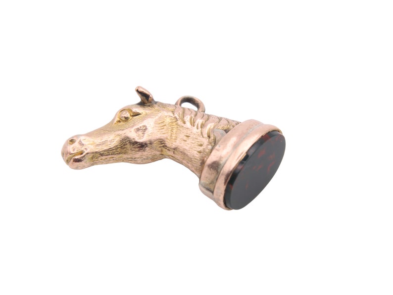 May include: A gold-colored horse head pendant with a dark red, circular stone base. The horse head is detailed with a textured mane and facial features. The pendant has a small loop at the top for hanging.