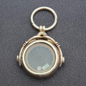 K&ouml;nnte beinhalten: Ein goldfarbener, runder Anh&auml;nger mit einem durchsichtigen Glaskern, der ein dekoratives Design aufweist. Der Anh&auml;nger hat eine Scharnier&ouml;se und einen kleinen Ring zur Befestigung. Der Hintergrund ist eine dunkle, strukturierte Oberfl&auml;che.