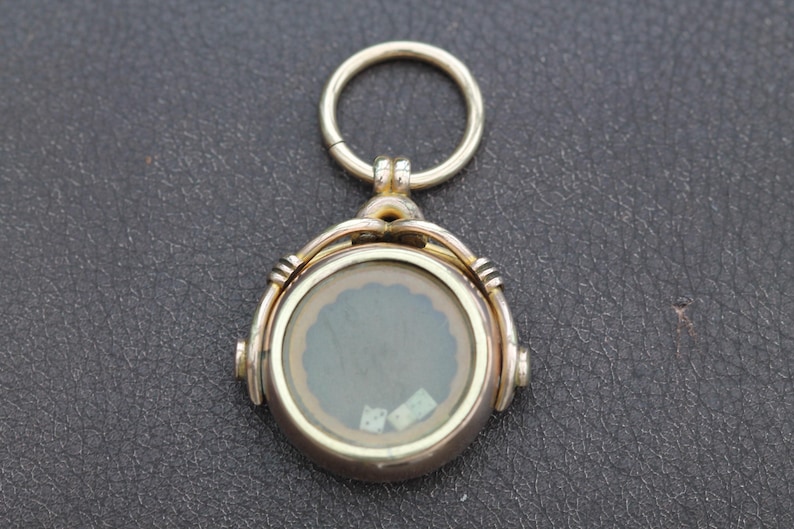 K&ouml;nnte beinhalten: Ein goldfarbener, runder Anh&auml;nger mit einem durchsichtigen Glaskern, der ein dekoratives Design aufweist. Der Anh&auml;nger hat eine Scharnier&ouml;se und einen kleinen Ring zur Befestigung. Der Hintergrund ist eine dunkle, strukturierte Oberfl&auml;che.