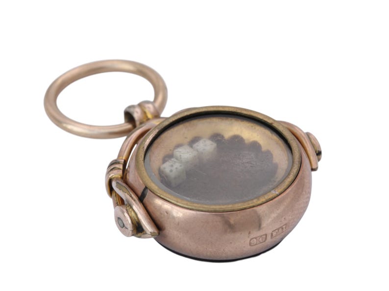 K&ouml;nnte beinhalten: Ein Vintage-Anh&auml;nger in Goldoptik mit einer transparenten, kreisf&ouml;rmigen Oberfl&auml;che. Der Anh&auml;nger hat oben eine Schlaufe und einen kleinen, runden K&ouml;rper. Im Inneren der Oberfl&auml;che befinden sich kleine, wei&szlig;e Objekte. Der Anh&auml;nger ist wahrscheinlich ein Charm oder ein Medaillon.
