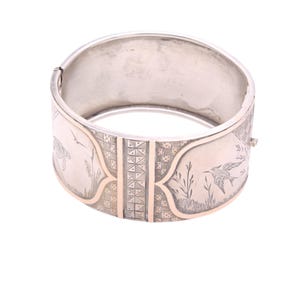 Könnte beinhalten: Silbernes, aufklappbares Armreif-Armband mit goldenen Akzenten. Das Armband hat eingravierte Vogel- und Pflanzenmotive. Die goldenen Akzente befinden sich an den Seiten und in der Mitte. Das Armband ist offen.