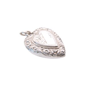 May include: A silver heart-shaped pendant with intricate floral and scrollwork detailing. The pendant has a small loop at the top for a chain and a blank, rectangular space in the center. The locket is on a white background.