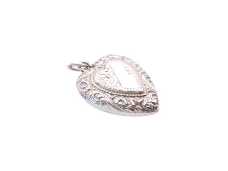 May include: A silver heart-shaped pendant with intricate floral and scrollwork detailing. The pendant has a small loop at the top for a chain and a blank, rectangular space in the center. The locket is on a white background.