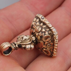 May include: A gold-colored pendant with intricate floral detailing. The pendant features a loop for hanging and a detailed floral design on the main body. The design includes various flowers and leaves, creating a vintage aesthetic.