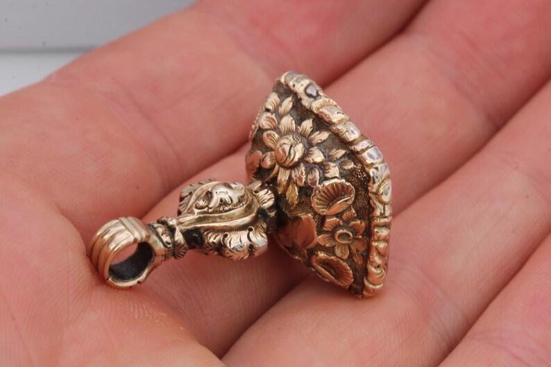 May include: A gold-colored pendant with intricate floral detailing. The pendant features a loop for hanging and a detailed floral design on the main body. The design includes various flowers and leaves, creating a vintage aesthetic.